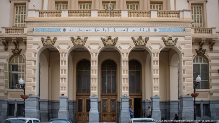 Arcaded entrance of Constantine city hall with tall wooden doors, columns, balconies, and Arabic inscription above.