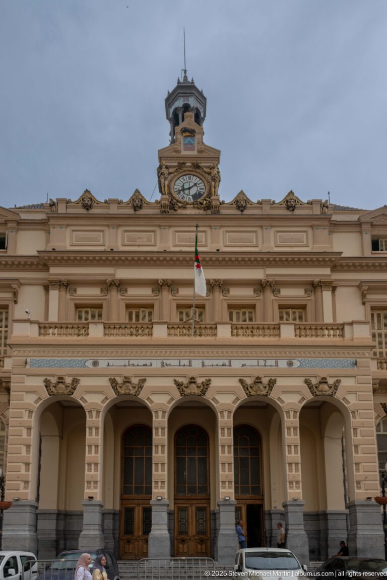 Frontal view of Constantine city hall with clock tower, Algerian flag, Arabic inscription, balcony, and arched entrance.