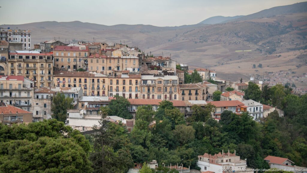 Dense Constantine hillside neighborhood with multi-story buildings, trees, and dry mountains rising in the background.