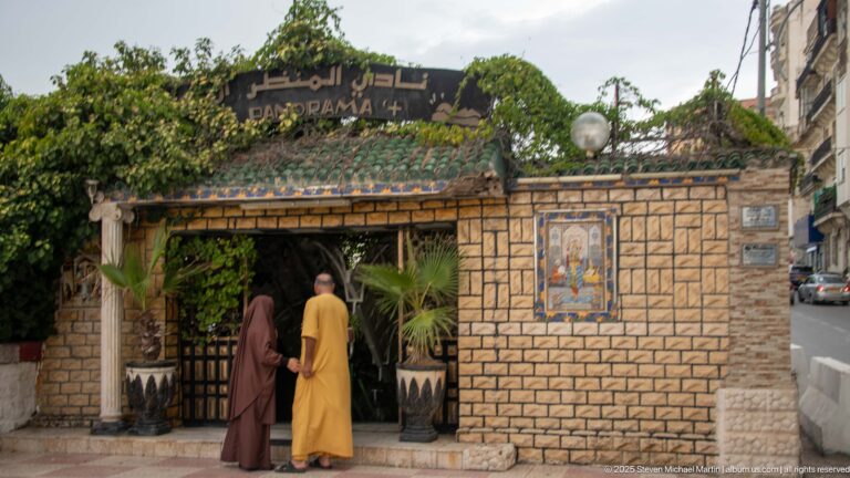 Two people entering a restaurant with Arabic signage, tiled canopy, stone-patterned walls, plants, and decorative ceramic panel.