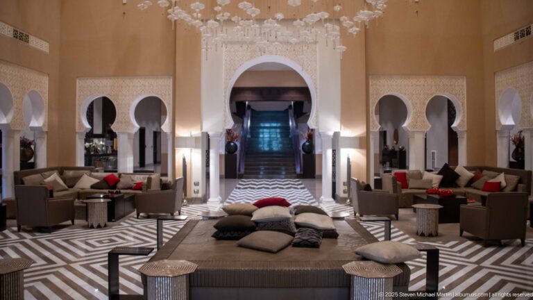 Symmetrical hotel lounge with horseshoe arches, carved screens, low seating, black-and-white geometric flooring, and a mirrored ceiling feature.