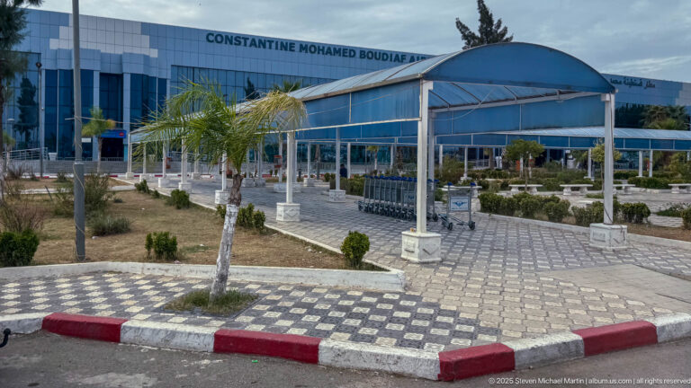 Constantine Mohamed Boudiaf Airport terminal with blue glazing, curved roof, palms, patterned paving, and covered walkway.