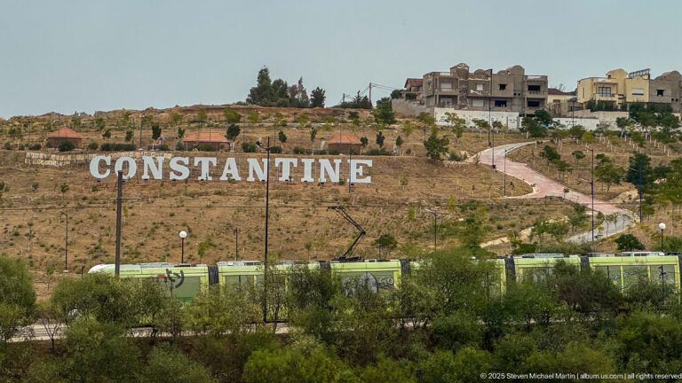 Large white CONSTANTINE hillside letters above a green tram, with a winding path and houses on the ridge.