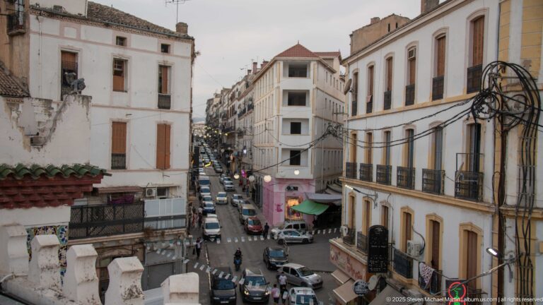 Narrow descending Constantine street lined with old multi-story buildings, balconies, cables, cars, awnings, and pedestrians.