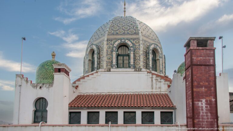 Frontal view of large patterned tiled dome above red roof and white walls, flanked by smaller green domes.