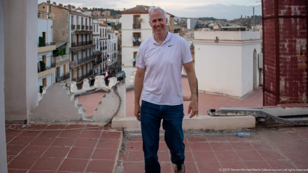 Man in a white polo shirt standing on a red-tiled rooftop terrace with Constantine buildings behind him.