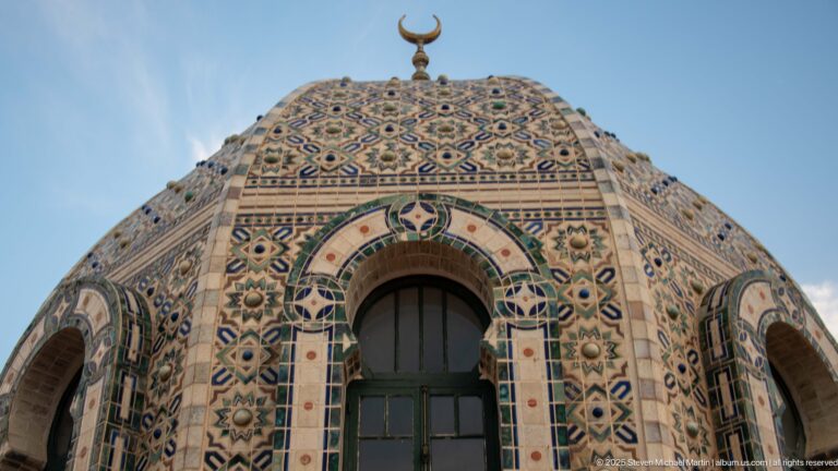 Low-angle close view of colorful geometric tiled dome with tall arched window and crescent finial.