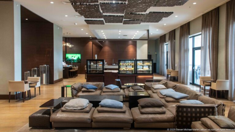 Modern hotel lounge with clustered sofas, café tables, display cases, tall windows, and suspended decorative ceiling panels.