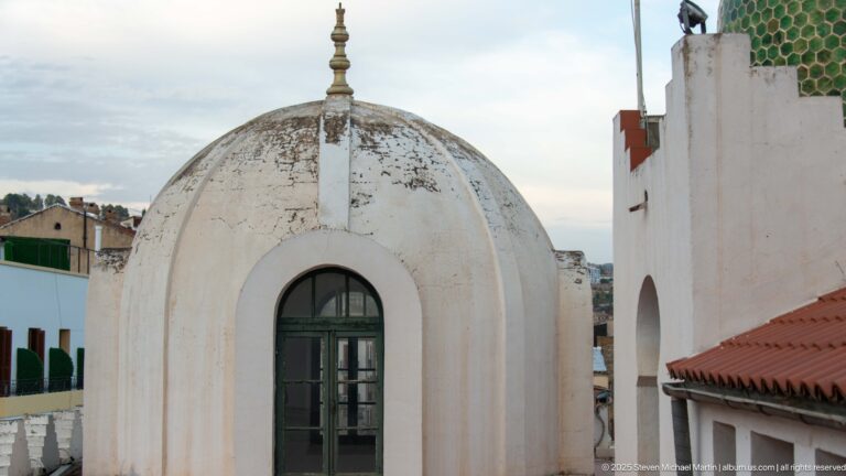 Small weathered white dome with tall arched window and finial beside a green-tiled dome and red roof.