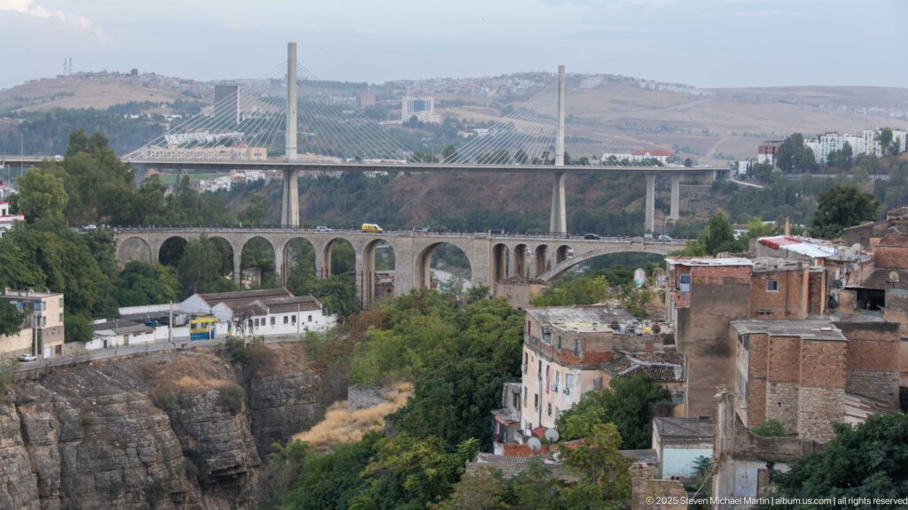 Modern cable-stayed bridge behind an older multi-arch viaduct across Constantine’s rocky valley and urban outskirts.
