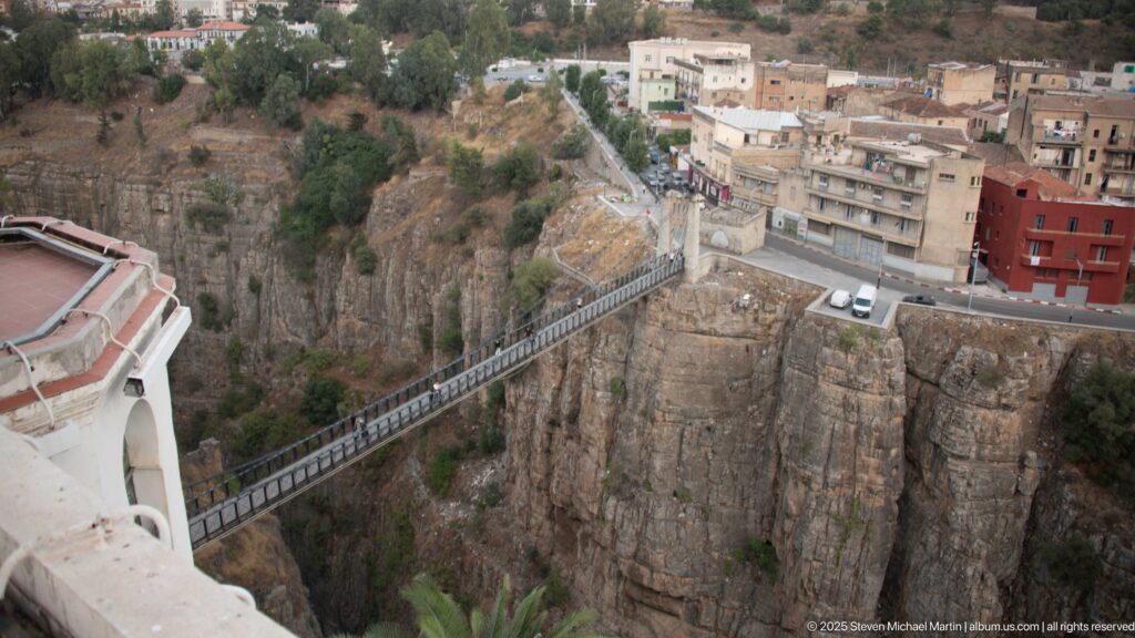 Narrow metal pedestrian bridge spanning sheer Constantine gorge cliffs between buildings and a road.