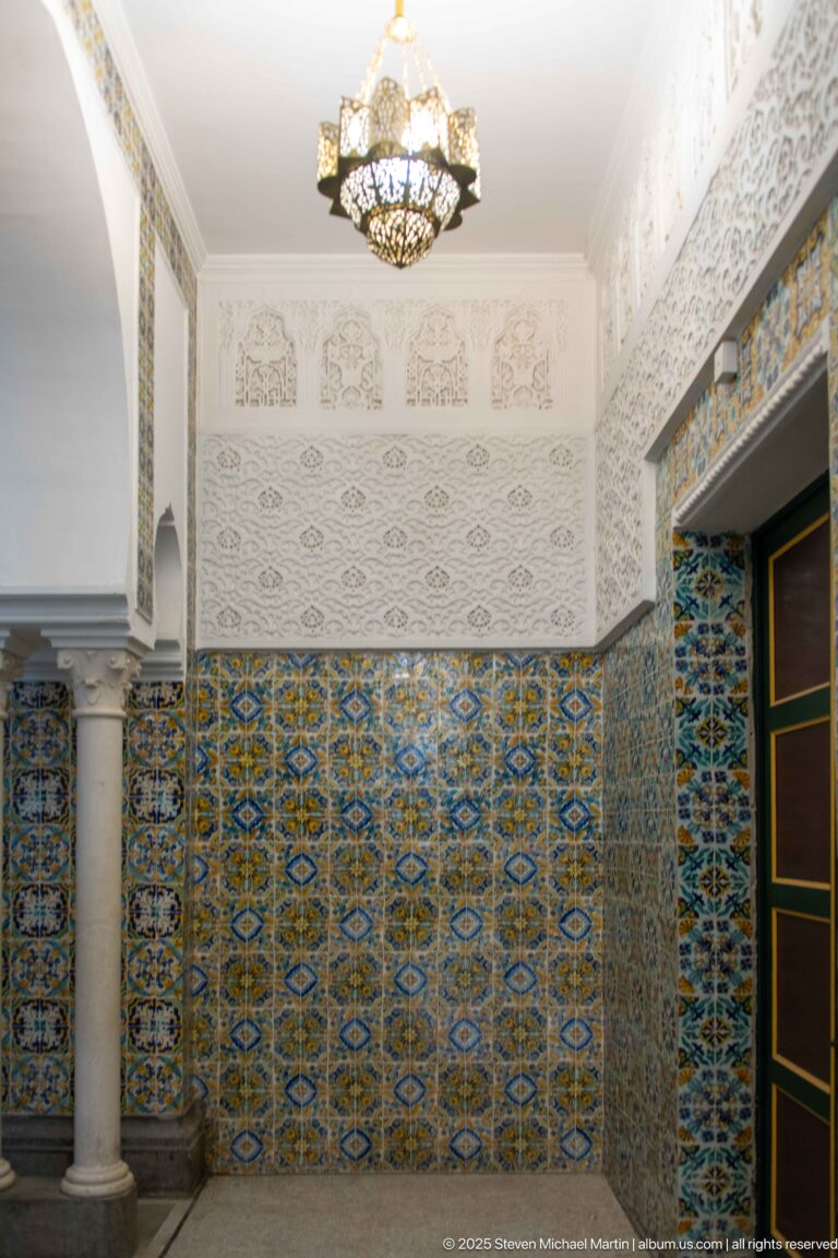 Interior with blue patterned tile walls, carved white plaster panels, a slender column, dark door, and hanging lantern.