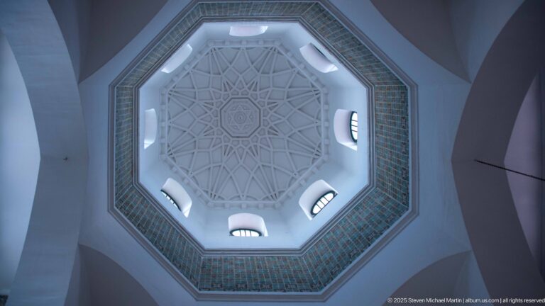 Upward view of an octagonal white geometric ceiling ringed by small arched windows and a band of patterned tiles.