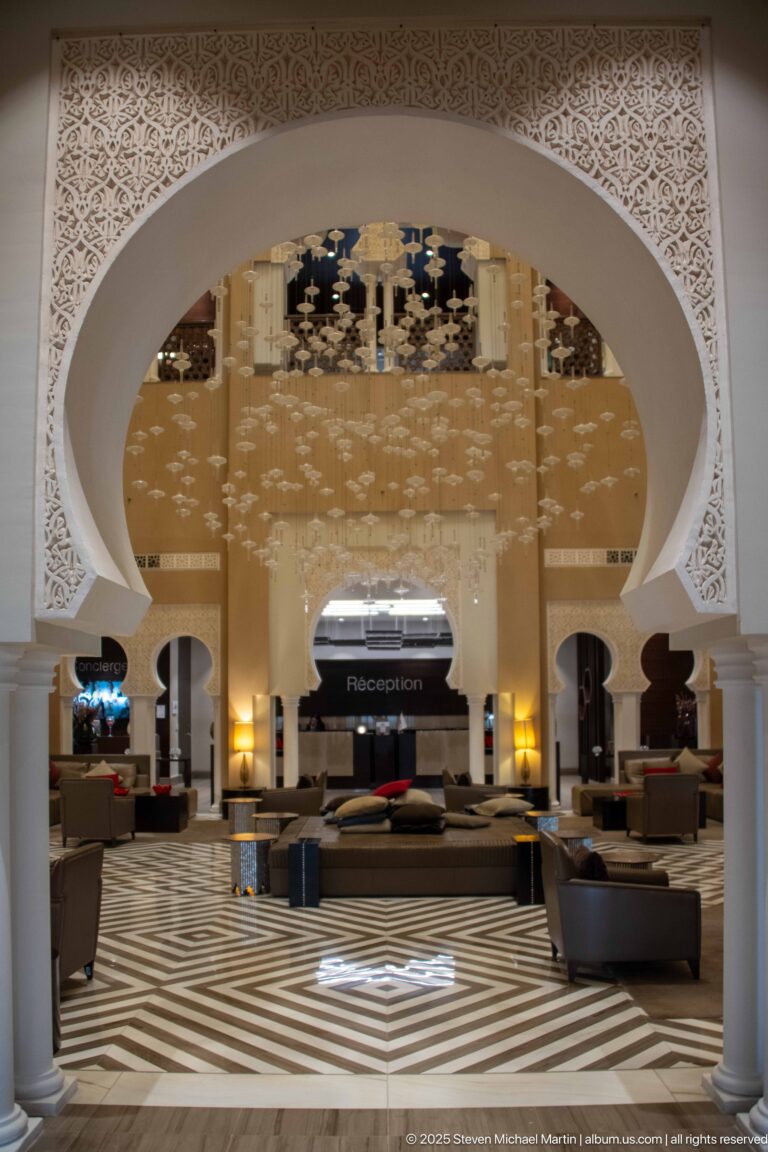 Hotel reception framed by a large horseshoe arch, white pierced ornament, mirrored ceiling, striped floor, and lounge seating.