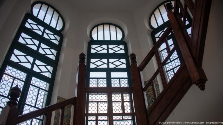 Tall geometric-glazed windows behind a dark wooden staircase in a bright historic interior.