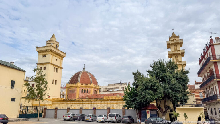 Mosque with a red ribbed dome and two pale minarets rising behind parked vehicles and trees in Constantine.