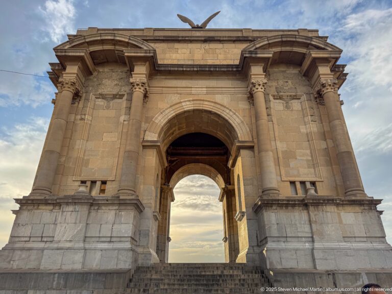 Stone triumphal-arch war memorial with broad steps, classical columns, and a winged figure on top against the sky.