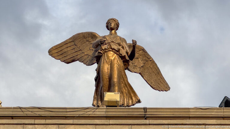 Bronze-colored winged female statue with outstretched wings standing above the Monument aux Morts against a cloudy sky.