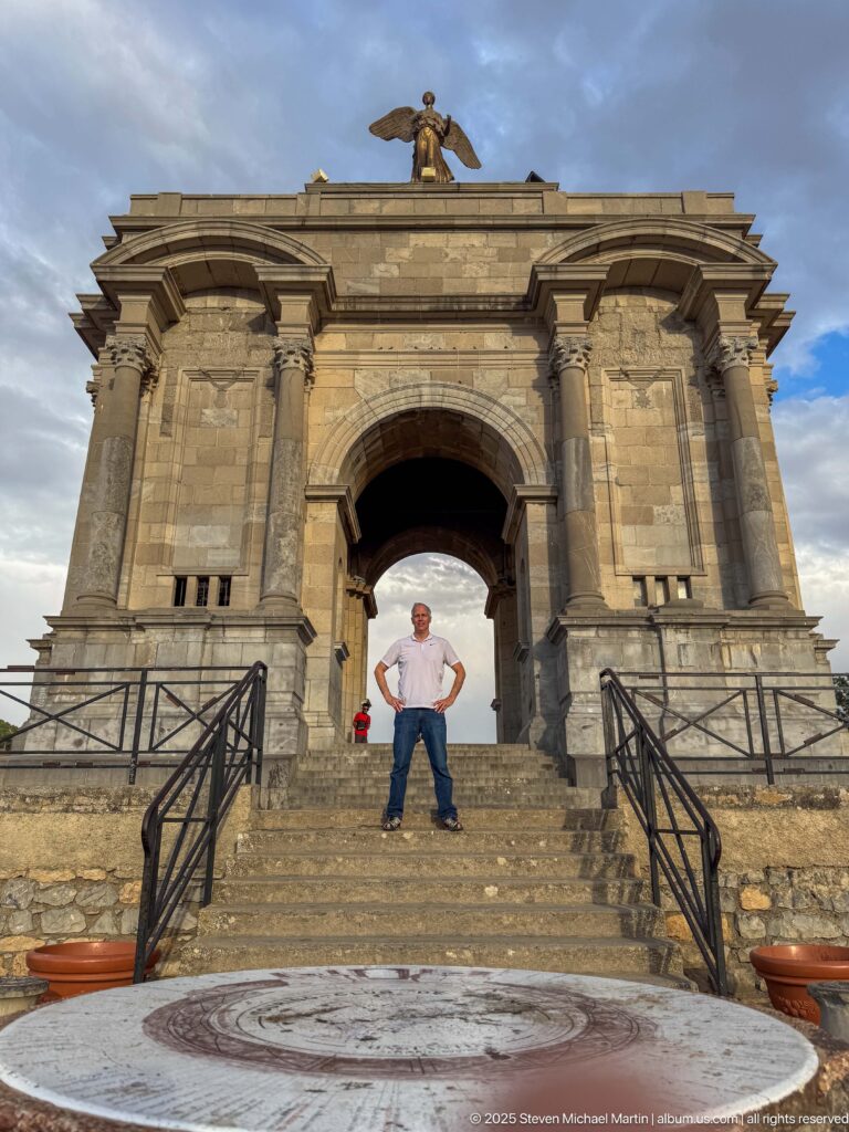 Visitor standing on the steps beneath Constantine’s massive stone Monument aux Morts arch, with a winged figure above.