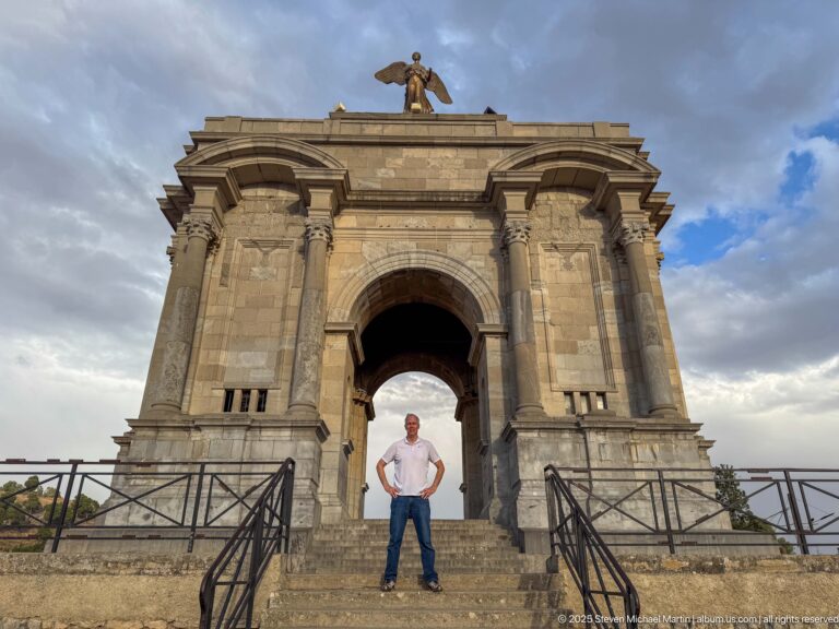 Visitor centered beneath Constantine’s Monument aux Morts, framed by the stone arch and winged figure above.