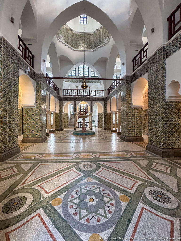 Wide tiled hall with white arches, upper galleries, geometric mosaic floor panels, hanging lantern, and central fountain-like feature.