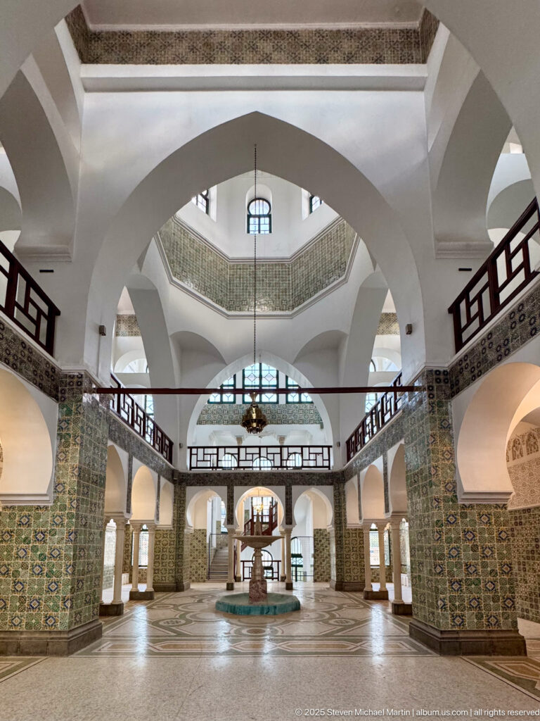 Symmetrical arched hall with tiled walls, dark upper railings, hanging lantern, and a central fountain-like feature.