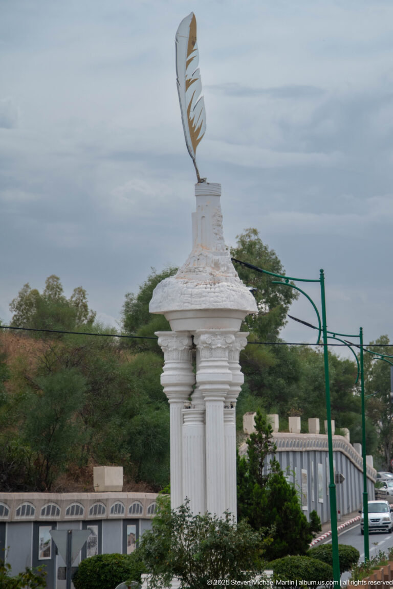 White column-like urban monument topped by a large stylized feather, with roadway structures and trees behind it.