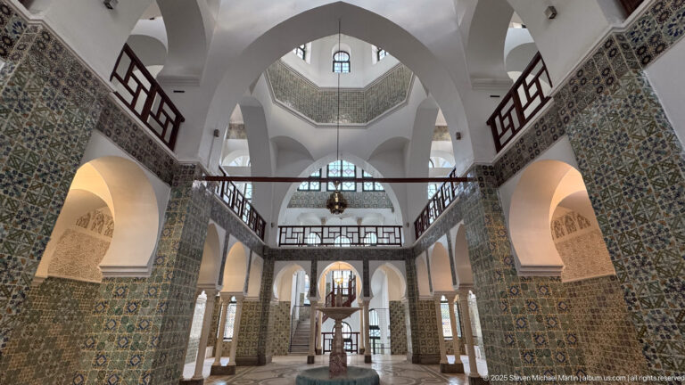 Tall white arches rising above tiled piers and a dark wood gallery, with a central lantern and fountain-like feature beyond.