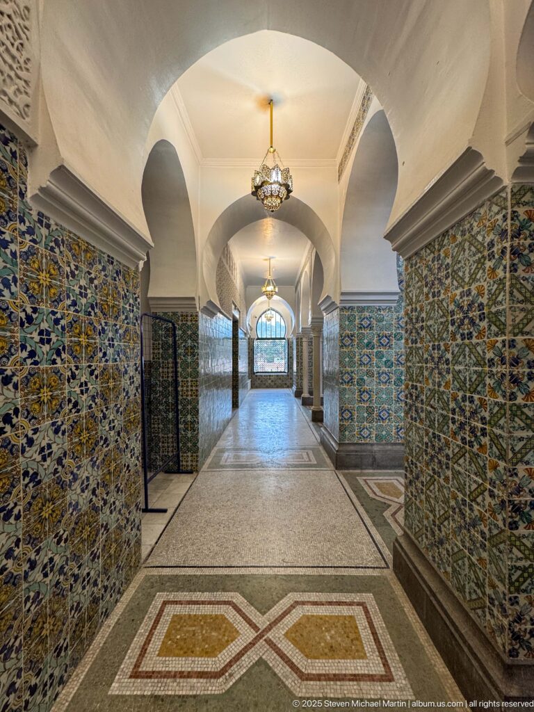 Long vaulted corridor with patterned tiled walls, hanging lanterns, geometric floor panels, and a bright arched window at the end.