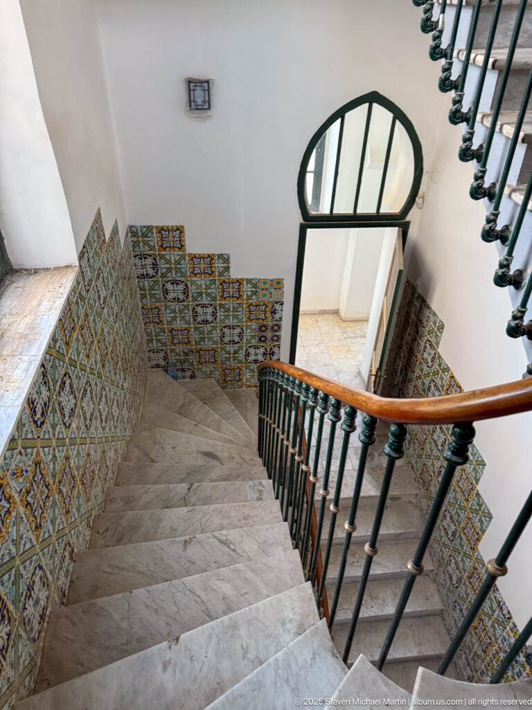 Marble staircase with dark green railing and wood handrail turning between walls lined with patterned ceramic tiles.