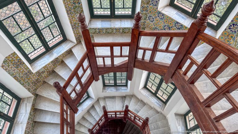 Top-down view of a multi-level stairwell with dark wooden railings, pale stone steps, tiled walls, and tall windows.