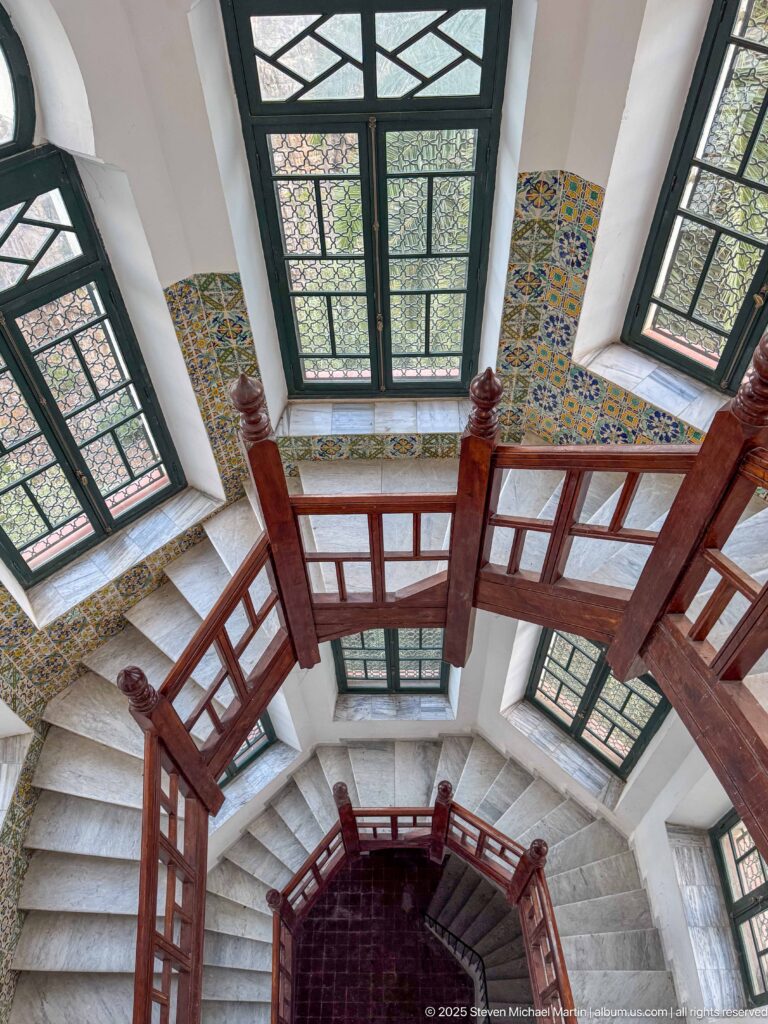 Oblique view down a wooden staircase with tall dark-framed windows, patterned wall tiles, and pale stone steps.