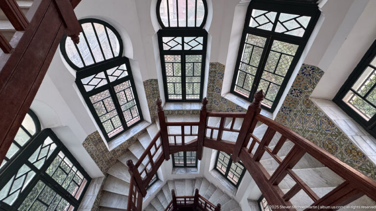 Steep view through a vertical stairwell shaft framed by dark wooden rails, tall windows, white walls, and patterned tiles.