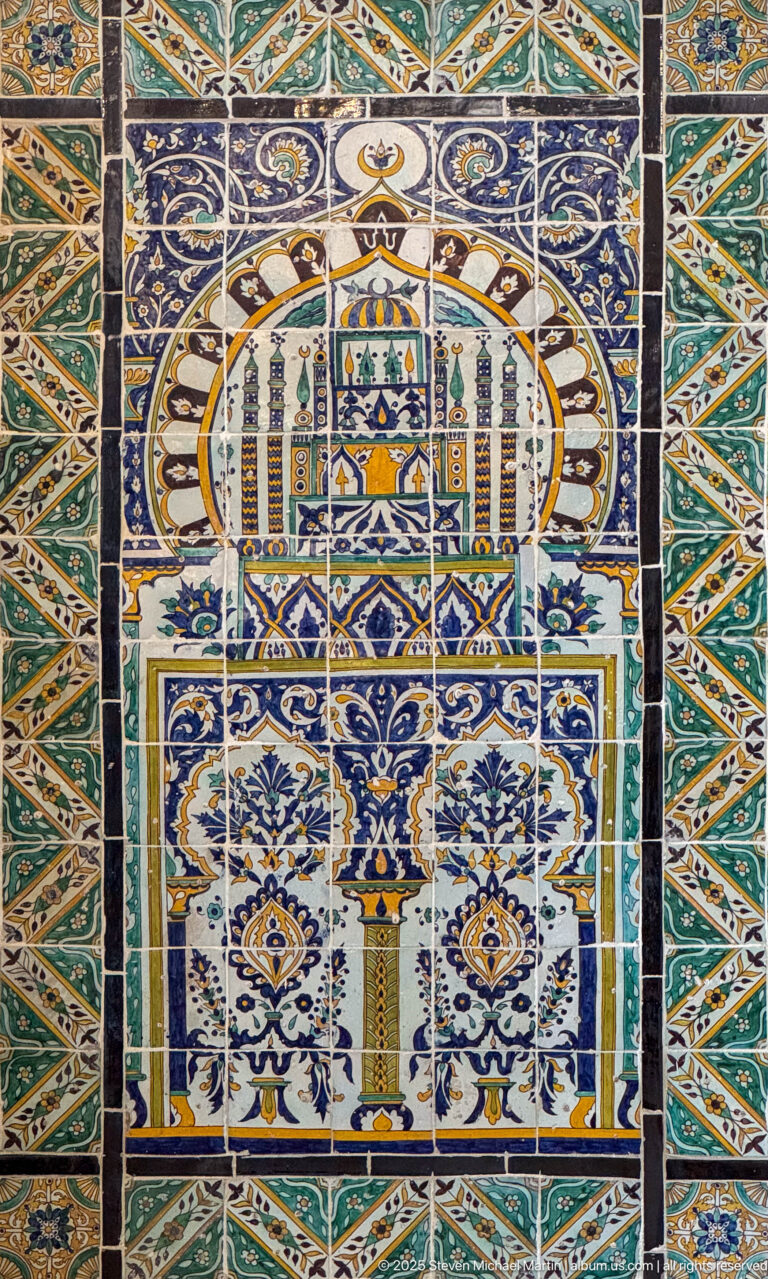 Polychrome ceramic tile panel with a stylized architectural scene framed by dense floral and geometric borders.