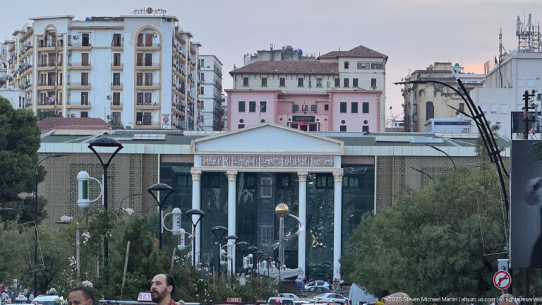 Modern institutional building with tall entrance columns and Arabic inscription, seen along a city street with apartments behind.