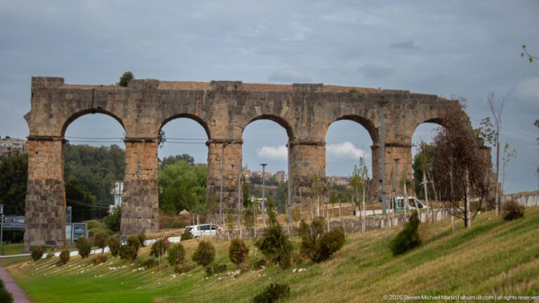 Weathered stone aqueduct with a row of tall semicircular arches beside a landscaped road in Constantine.