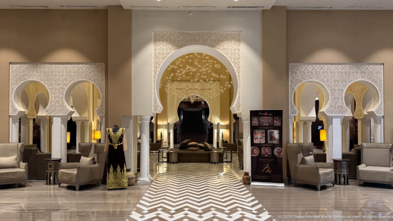 Symmetrical lobby with white horseshoe arches, slender columns, lounge seating, warm lamps, and a black-and-white chevron floor.