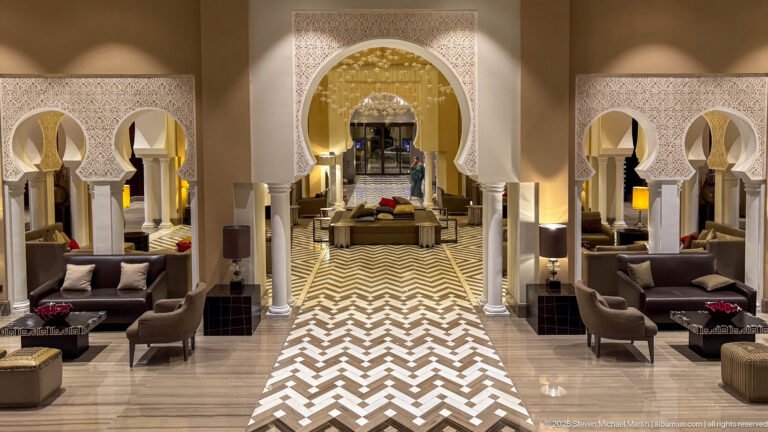 Long axial lobby view through white horseshoe arches with lounge seating, slender columns, and glossy chevron flooring.