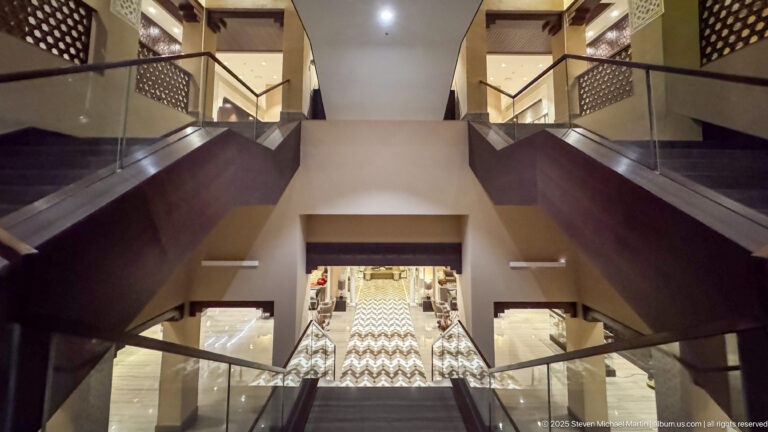 Symmetrical escalators and stairs inside a modern hotel, with glass railings, dark paneling, and a patterned lower-level corridor.