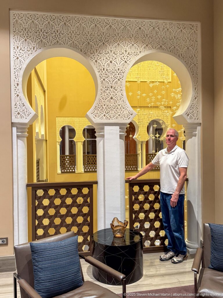 Hotel lounge with horseshoe arches, intricate geometric wall carving, warm gold lighting, low seating, and a standing visitor.