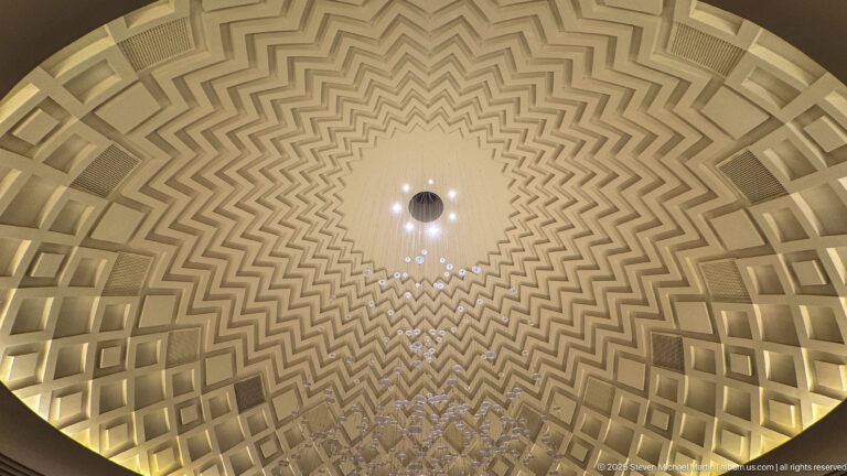 Upward view of a geometric coffered ceiling with stepped zigzag patterns, warm recessed lighting, and a central chandelier.