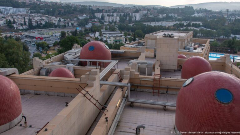 Flat hotel rooftops with several rounded red service structures, ladders, pipes, and Constantine’s buildings and hills beyond.