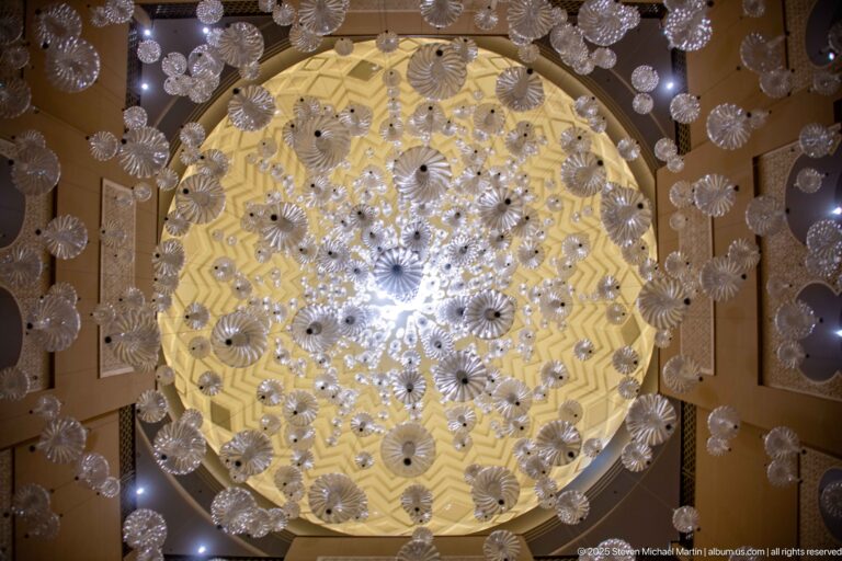 Large circular chandelier made of many clear flower-like glass elements suspended beneath a glowing geometric ceiling.
