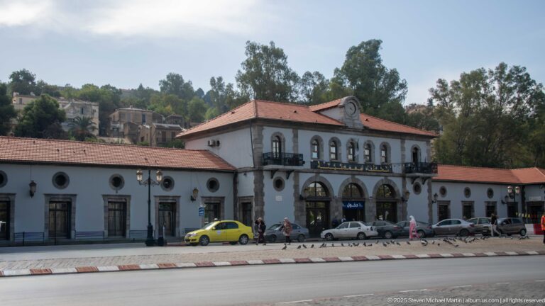 White transport building with a red-tiled roof, arched central bays, round windows, taxis, and pedestrians along the street.