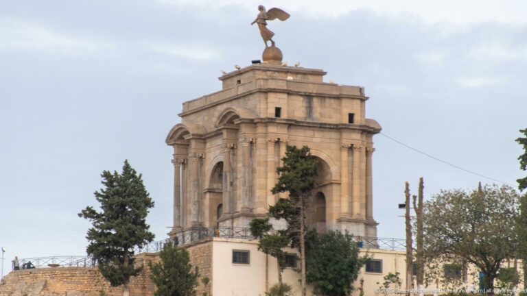 Stone triumphal-arch memorial topped by a winged figure, seen above trees in Constantine.