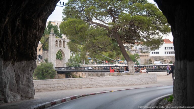 View from a rock-cut roadway toward a stone tower of Sidi M’Cid Bridge, with a large pine tree and pedestrians.