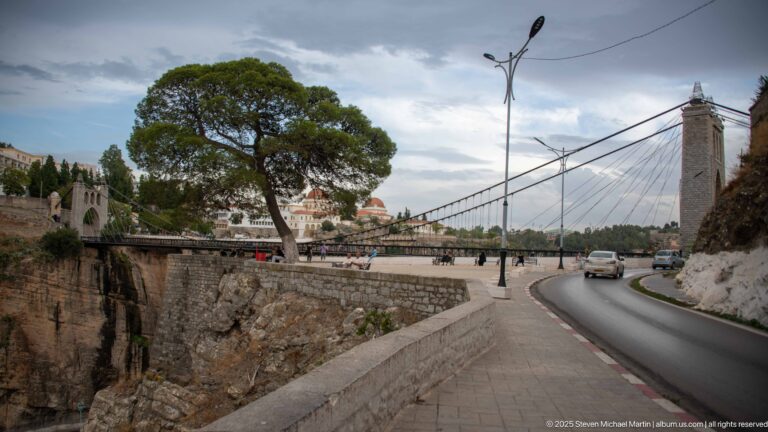 Sidi M’Cid suspension bridge beside a curving gorge road, with cars, pedestrians, and a stone tower.