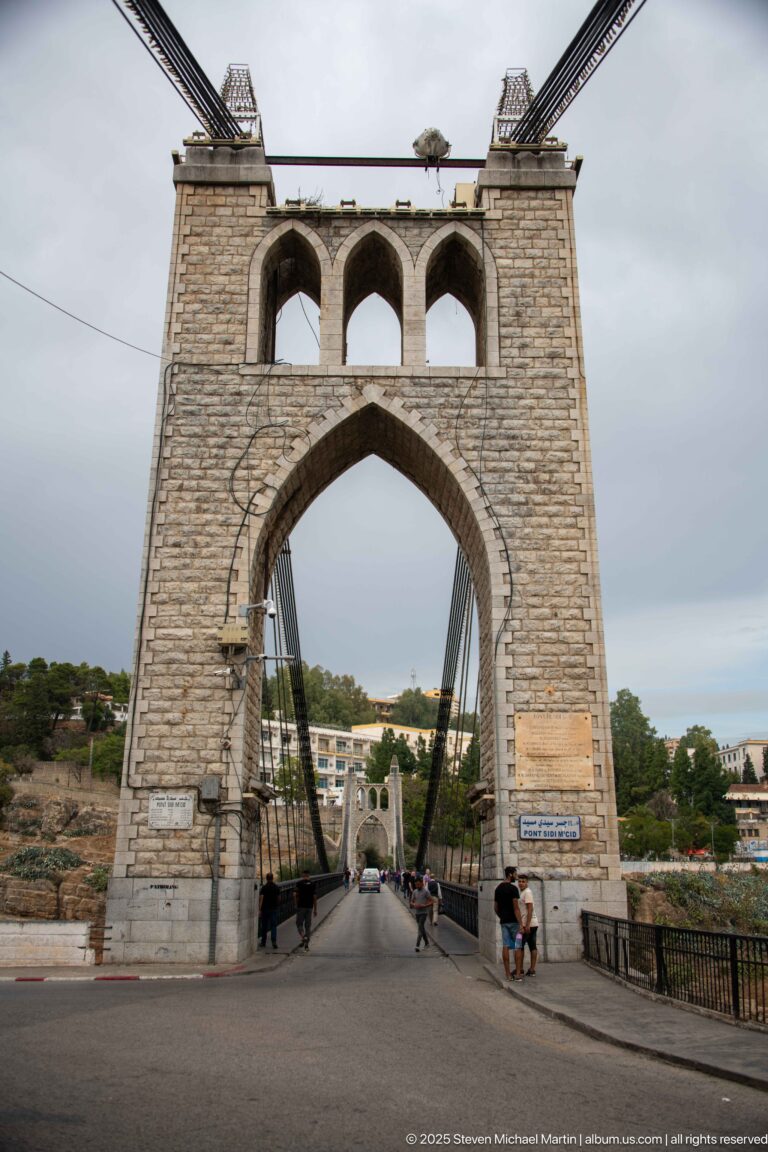 Roadway and pedestrians passing through the pointed stone portal of Sidi M’Cid Bridge beneath suspension cables.