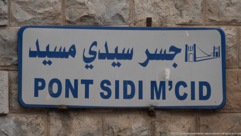 Bilingual blue-and-white sign on stone wall reading “PONT SIDI M’CID” in Arabic and Latin script.