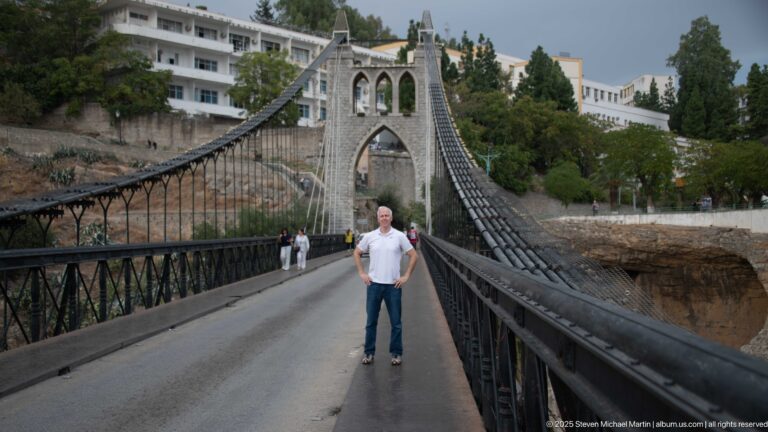 Man standing on the roadway of Sidi M’Cid Bridge, framed by suspension cables and the stone tower ahead.